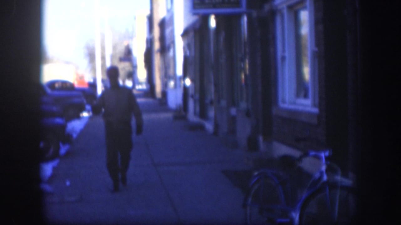 Man in dark clothing walks away from camera along a sidewalk flanked by mid-century storefronts and parked cars, strong blue color cast.