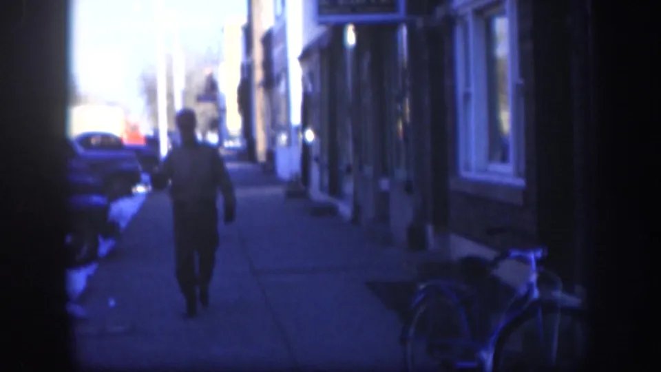 A man in dark clothing walks away from camera along a Kansas sidewalk circa 1958, with mid-century storefronts, late-1950s automobiles, and a bicycle visible to the right.