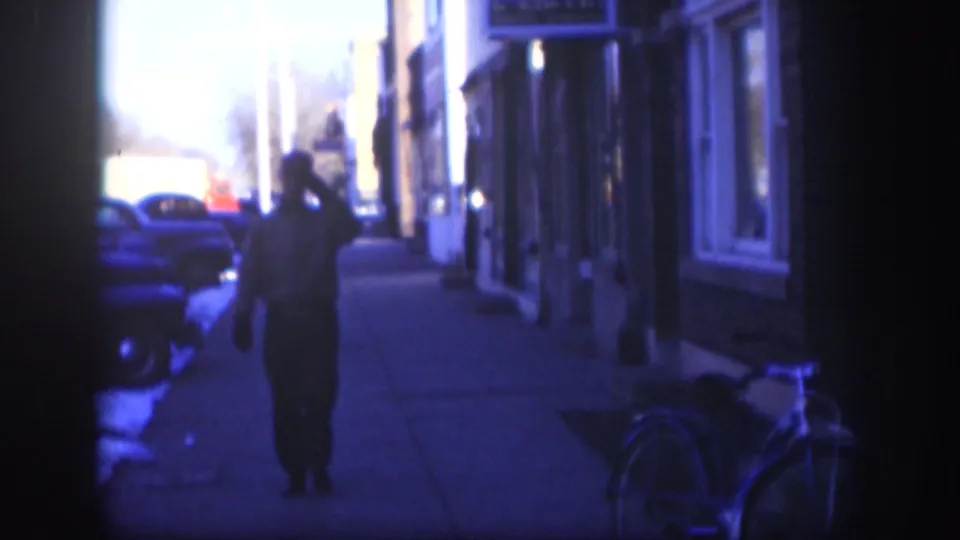 A man pauses on a Kansas main-street sidewalk circa 1958, his hand raised to his hat brim in a greeting gesture, with mid-century storefronts, automobiles, and a bicycle nearby.