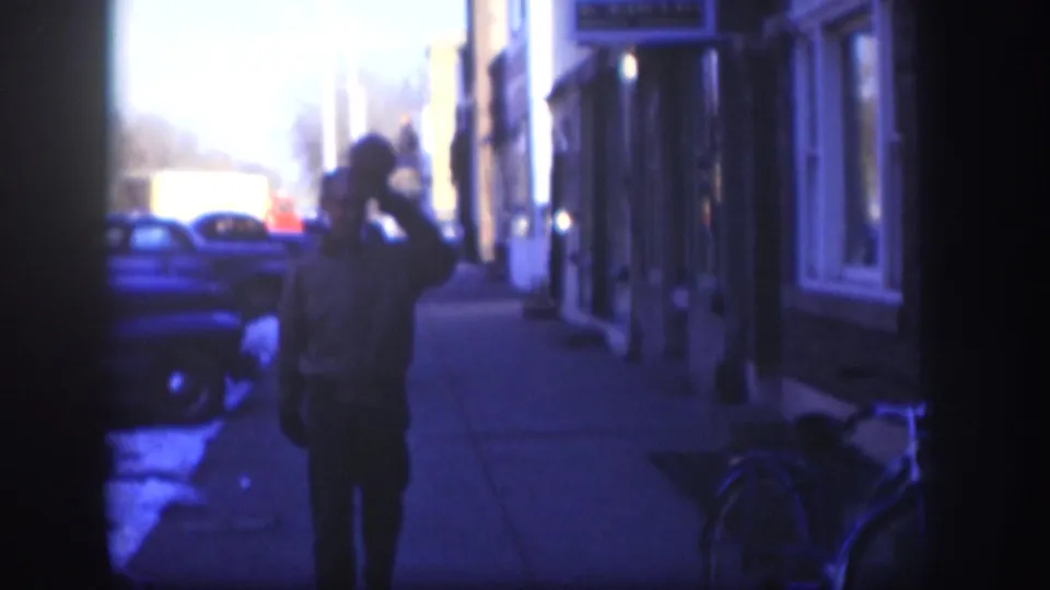Continuation of the hat-tip gesture: the man faces toward the camera on a Kansas sidewalk circa 1958, with storefronts, late-1950s automobiles, and a bicycle visible to the right.