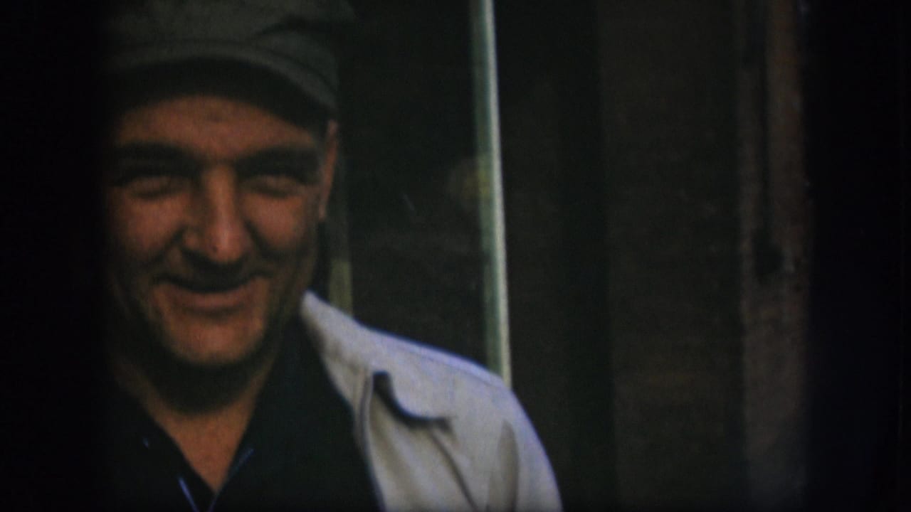 Extreme close-up of a middle-aged man in a flat cap looking slightly downward and smiling, 8mm film grain visible.