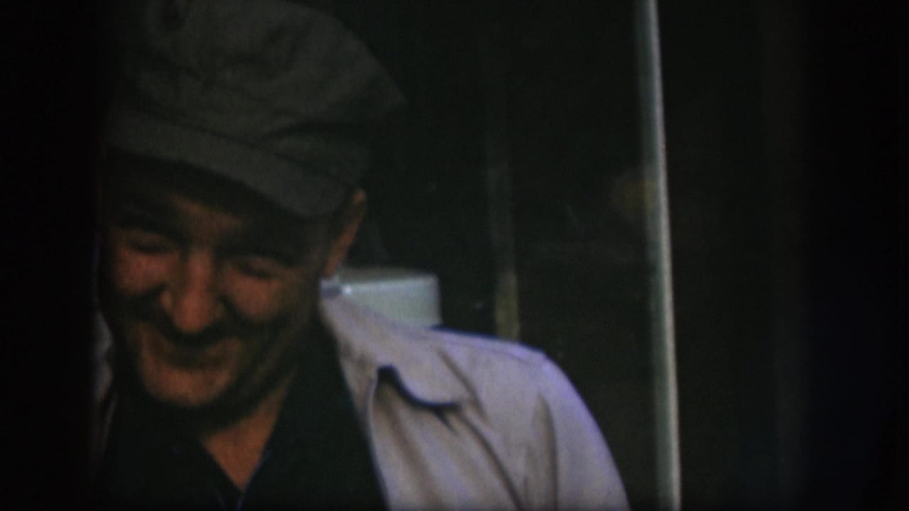 Man in flat cap and light jacket smiling broadly toward camera; a white appliance is partially visible at the right edge.