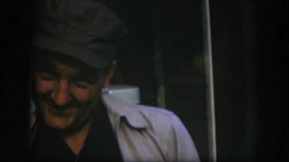 Middle-aged man in a flat cap speaks and smiles at the camera on 8mm film; a white appliance is partially visible at right, consistent with a 1958 Kansas laundry room.