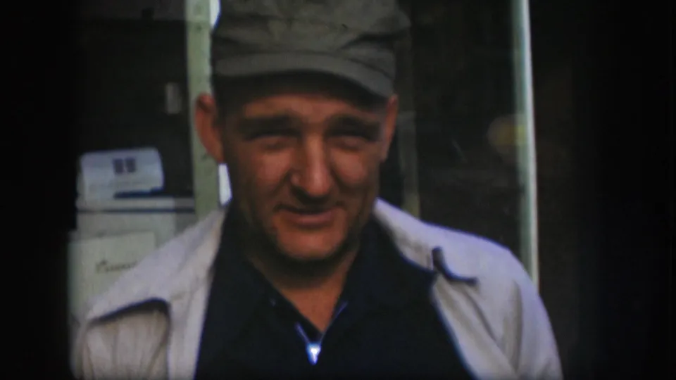 Middle-aged man in flat cap and light jacket filmed on 8mm in what appears to be a domestic laundry room circa 1958 Kansas; a white washing machine with a dark panel is visible behind him.