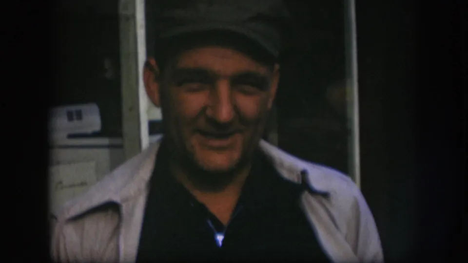 Close shot of a smiling middle-aged man in a flat cap, with a white domestic washing machine partially visible at right; 8mm home-movie footage consistent with late 1950s Kansas.