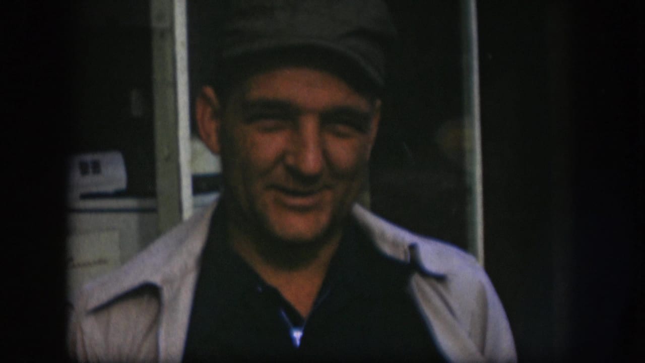Middle-aged man in grey flat cap and light open jacket smiles and speaks to camera in Kansas home movie footage, circa 1958.