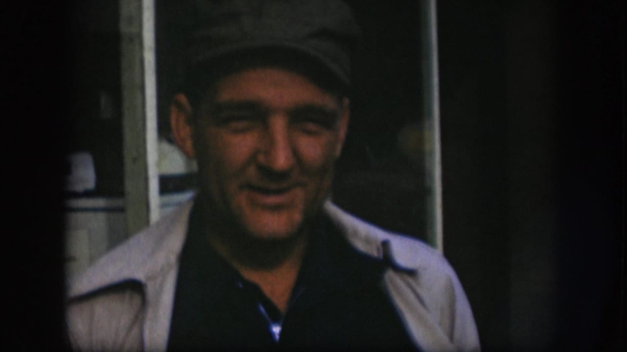 Man in grey flat cap and light jacket in Kansas home movie circa 1958, expression mid-shift as he continues speaking toward camera.