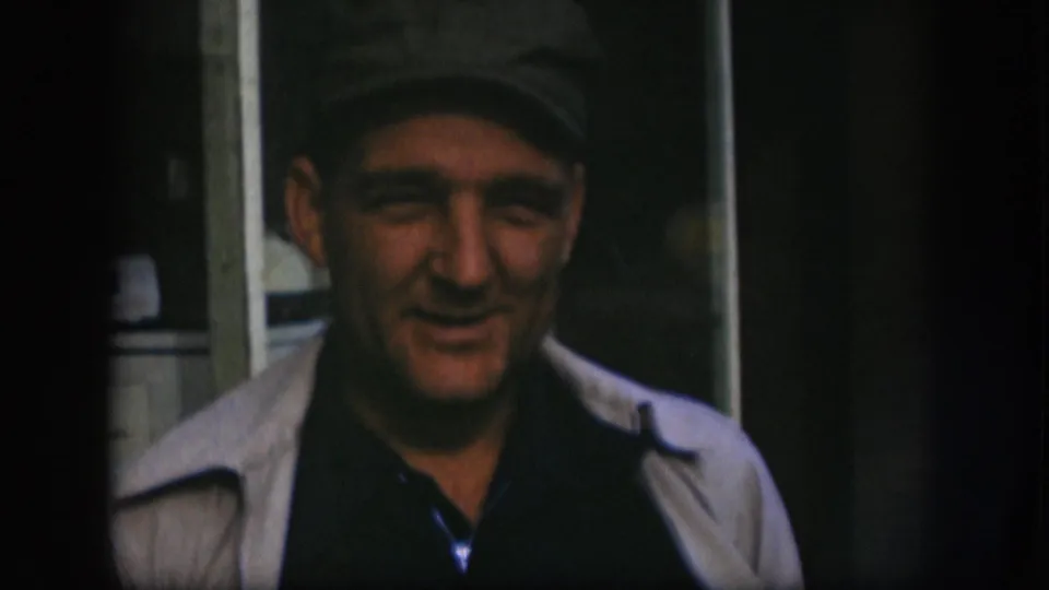 Man in grey flat cap and light jacket in Kansas home movie circa 1958, expression mid-shift as he continues speaking toward camera.