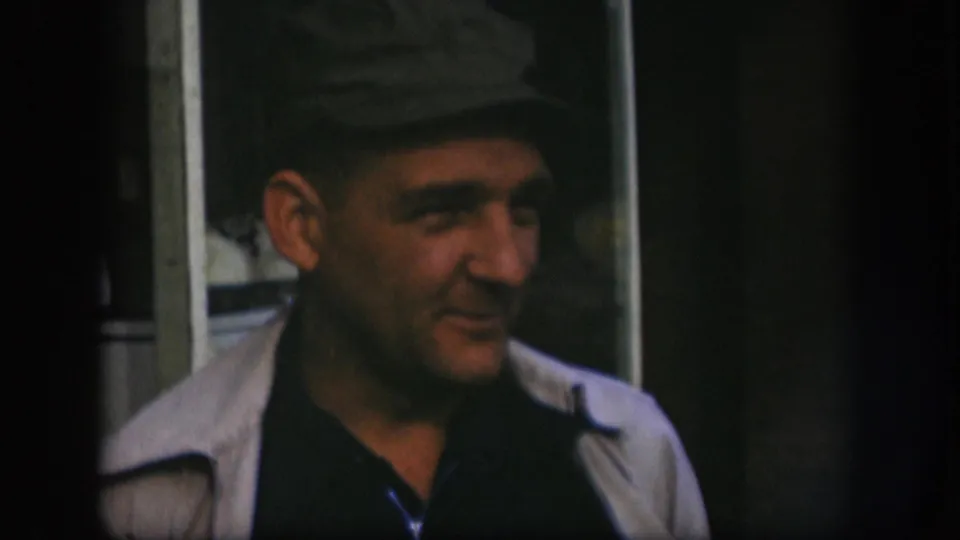 Man in grey flat cap turns to his left in Kansas home movie circa 1958, gaze moving off-camera in a candid conversational moment.