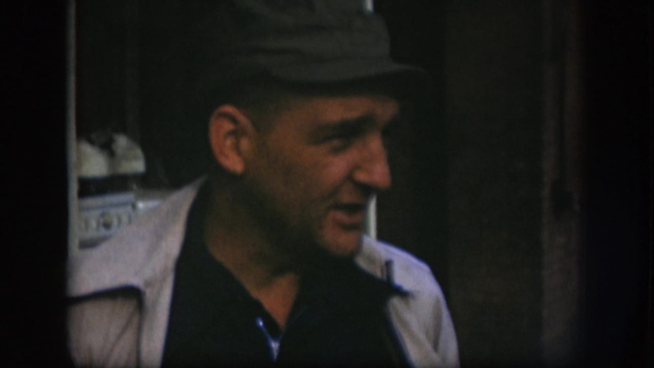Close-up of a middle-aged man in a flat dark cap and light jacket looking off-camera left, 8mm home-movie footage from Kansas, circa 1958.