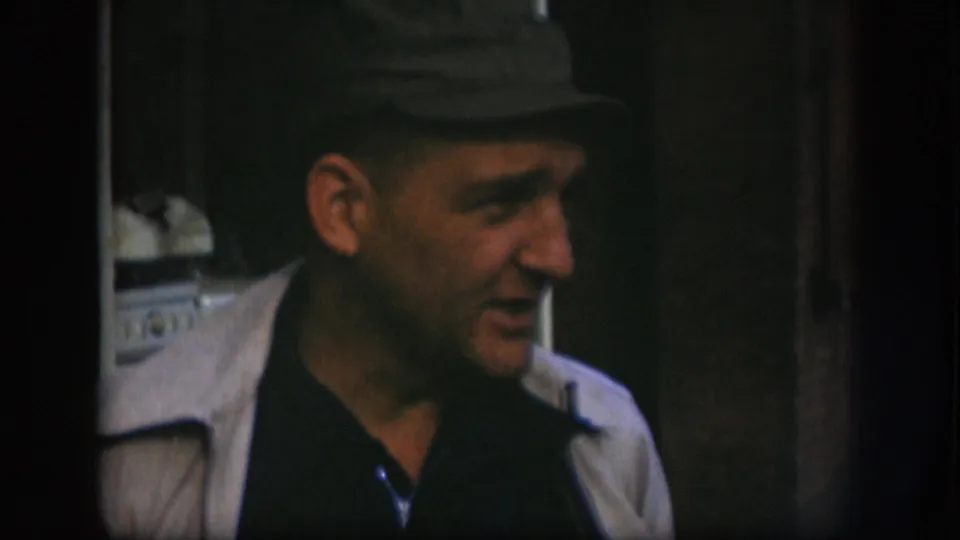 Close-up of a middle-aged man in a flat dark cap and light jacket looking off-camera left, 8mm home-movie footage from Kansas, circa 1958.