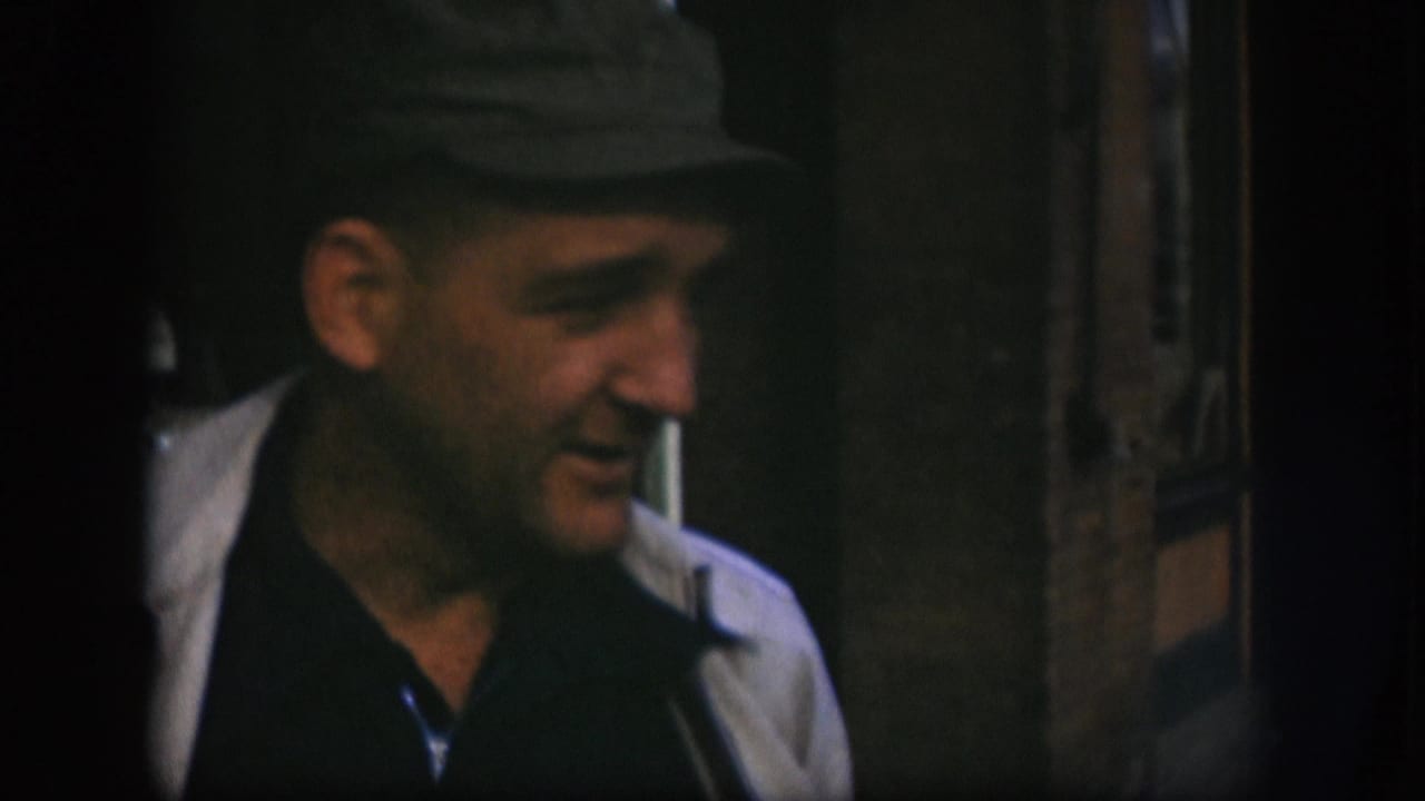 Man in flat dark cap and layered jacket facing right, possible cigarette at lips, wooden door frame visible at right edge.