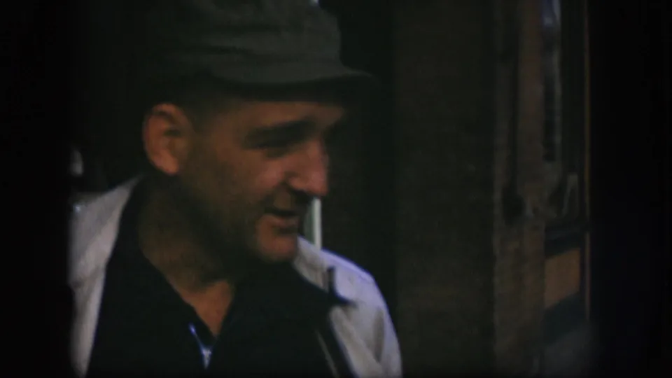 Close-up of man in dark flat cap facing right with a possible cigarette at his lips, wooden door frame at the frame edge, 8mm home-movie footage from Kansas, circa 1958.