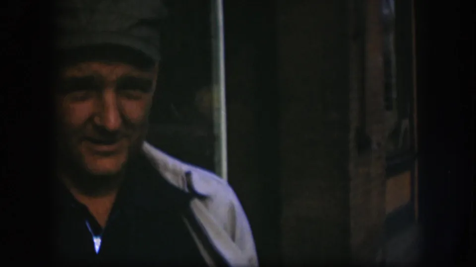 Close-up of a man in a flat dark cap beginning to turn toward camera, wearing a light jacket over a dark shirt, 8mm home-movie footage from Kansas, circa 1958.