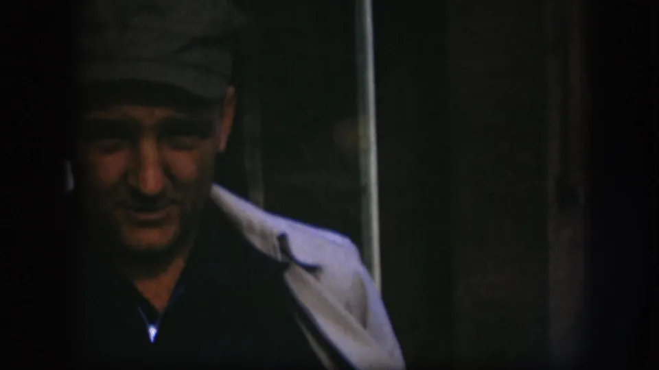 Close-up of a middle-aged man in a dark flat cap looking toward camera with a mild expression, dark doorway background, 8mm home-movie footage from Kansas, circa 1958.