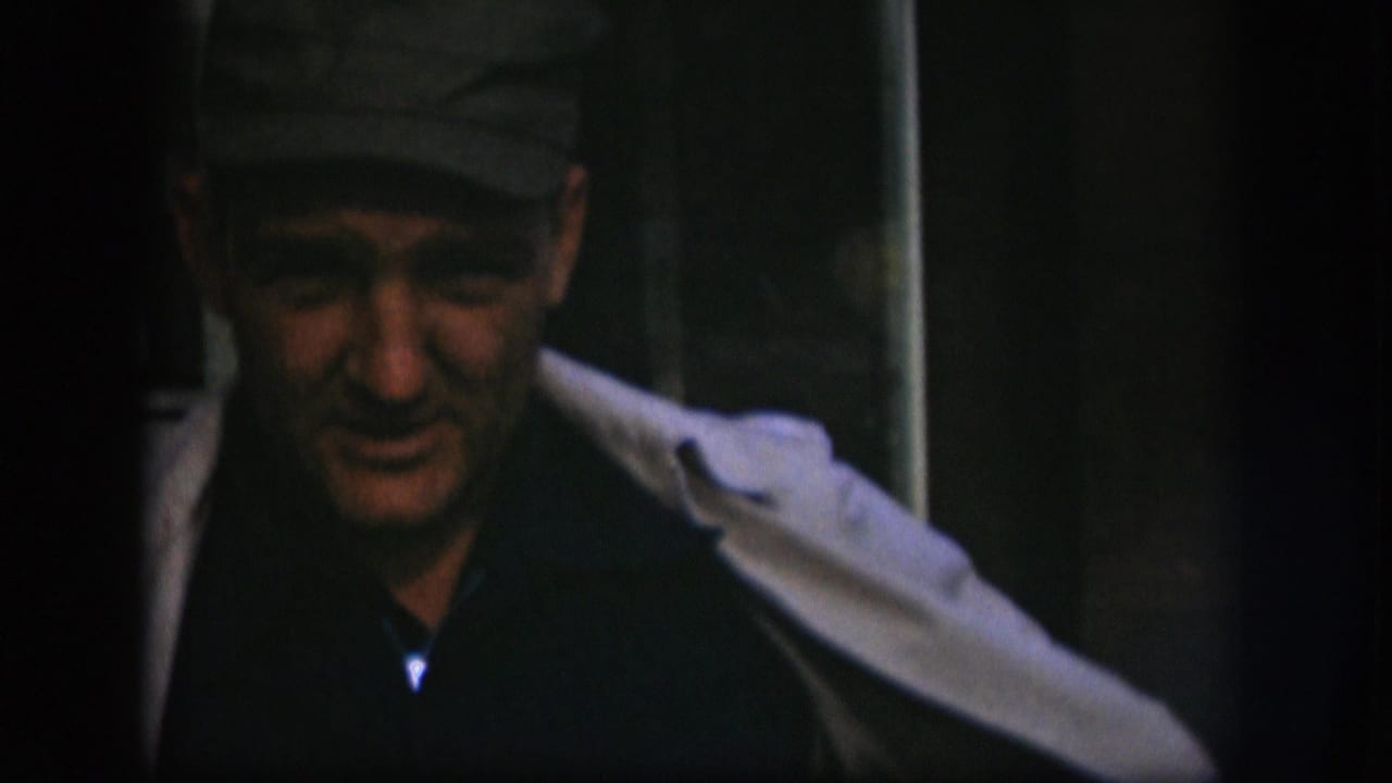 Middle-aged man wearing a dark flat cap and pale jacket over a dark shirt, looking toward the camera in what appears to be a 1958 Kansas 8mm home movie.