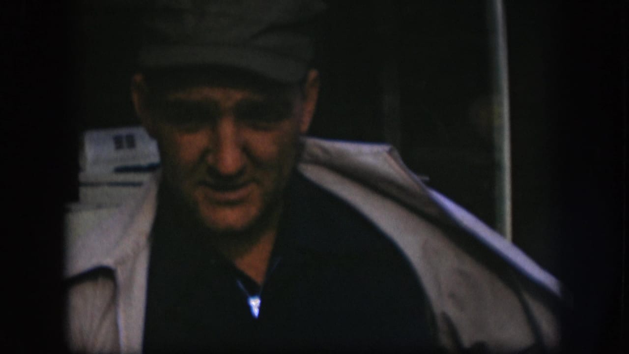 Close-up of man in dark flat cap appearing to speak, white appliance visible behind him; consistent with a 1958 Kansas home movie exterior setting.