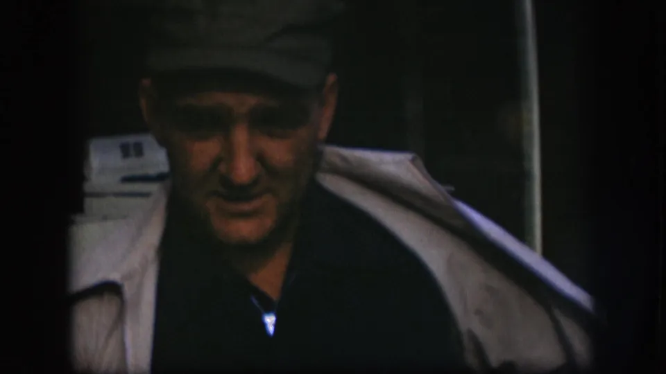 Close-up of man in dark flat cap appearing to speak, white appliance visible behind him; consistent with a 1958 Kansas home movie exterior setting.