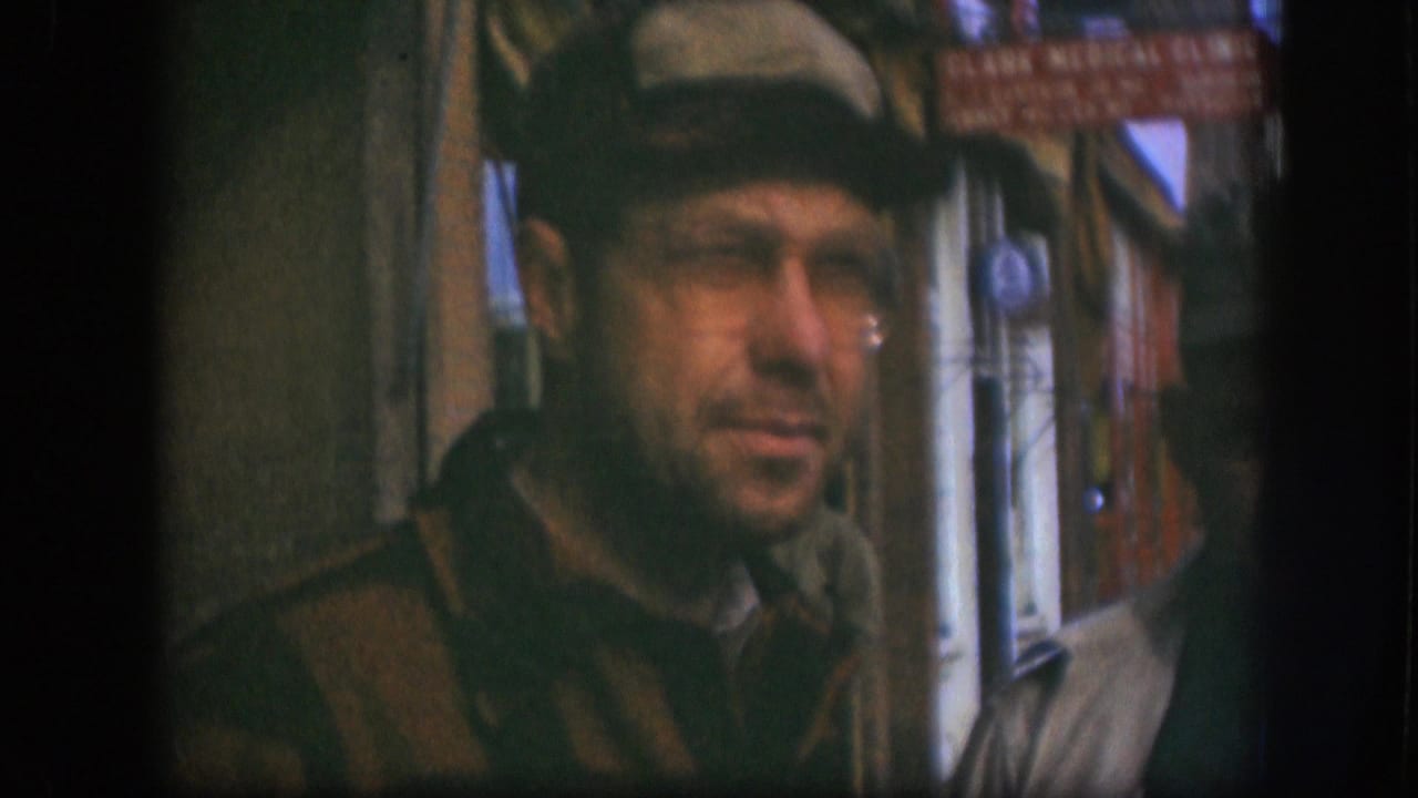 Close-up of a man in flat cap and wire-rimmed glasses on a Kansas city street, circa 1958; brick storefront and partial signage in background.