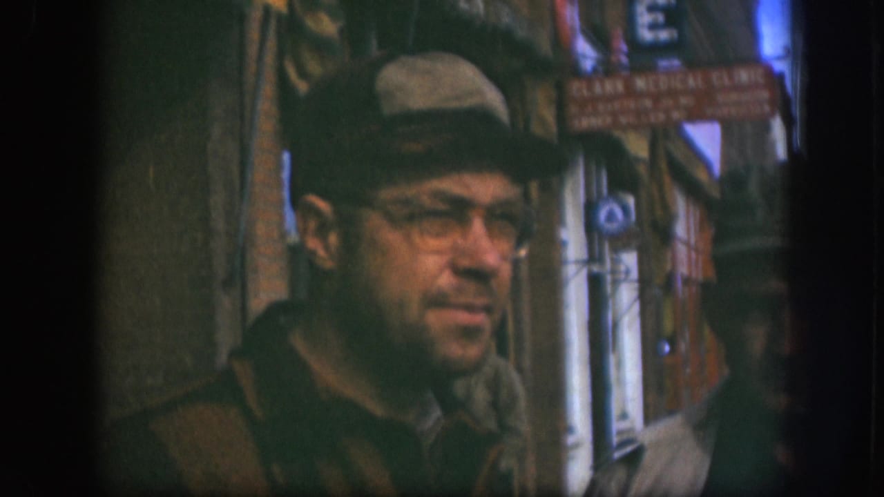 Man in flat cap and glasses in slight profile; Clark Medical Clinic sign legible on right; second person visible behind him.