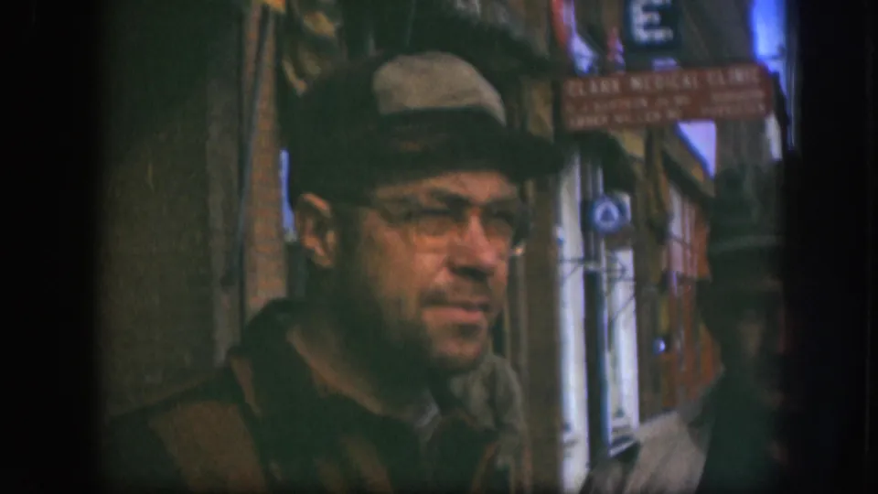 Medium close-up of man in flat cap, wire-rimmed glasses, and heavy coat on a Kansas sidewalk circa 1958, with Clark Medical Clinic sign readable in background.