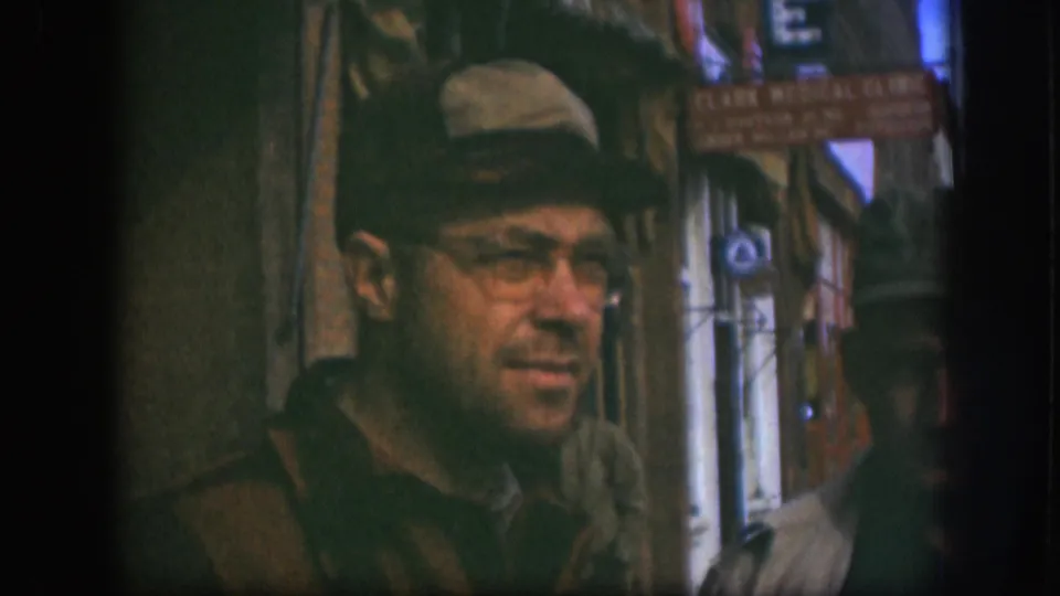 Close-up of man in flat cap and wire-rimmed glasses on a Kansas street circa 1958; Clark Medical Clinic sign at frame right, blue sky visible above storefront roofline.