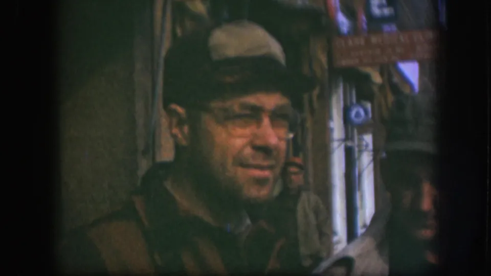Close-up of man in flat cap, wire-rimmed glasses, and dark coat on a Kansas street circa 1958; Clark Medical Clinic sign at upper right; another person at frame edge.