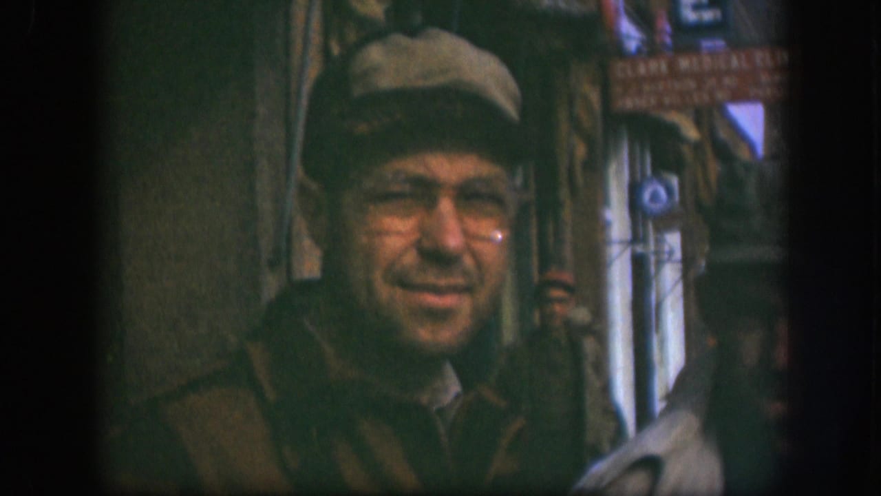 Man in newsboy cap and wire-rim glasses smiling toward camera outside a small shop in Kansas, USA, circa 1958; partial 'Clark Medical Clinic' sign legible at upper right.