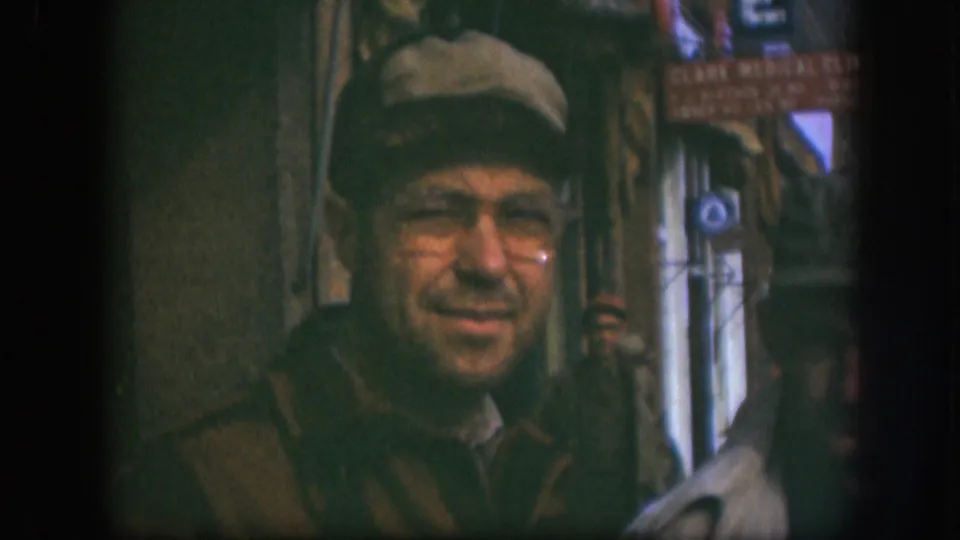 Man in newsboy cap and wire-rim glasses smiling toward camera outside a small shop in Kansas, USA, circa 1958; partial 'Clark Medical Clinic' sign legible at upper right.