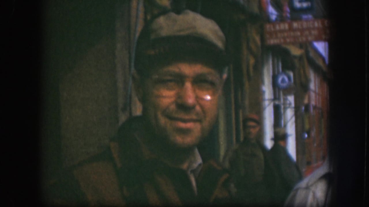 Close shot of a smiling man in newsboy cap and wire-rim glasses outside a Kansas storefront circa 1958; 'Clark Medical' lettering partially visible on a red sign behind him.