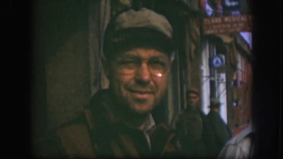 Close shot of a smiling man in newsboy cap and wire-rim glasses outside a Kansas storefront circa 1958; 'Clark Medical' lettering partially visible on a red sign behind him.