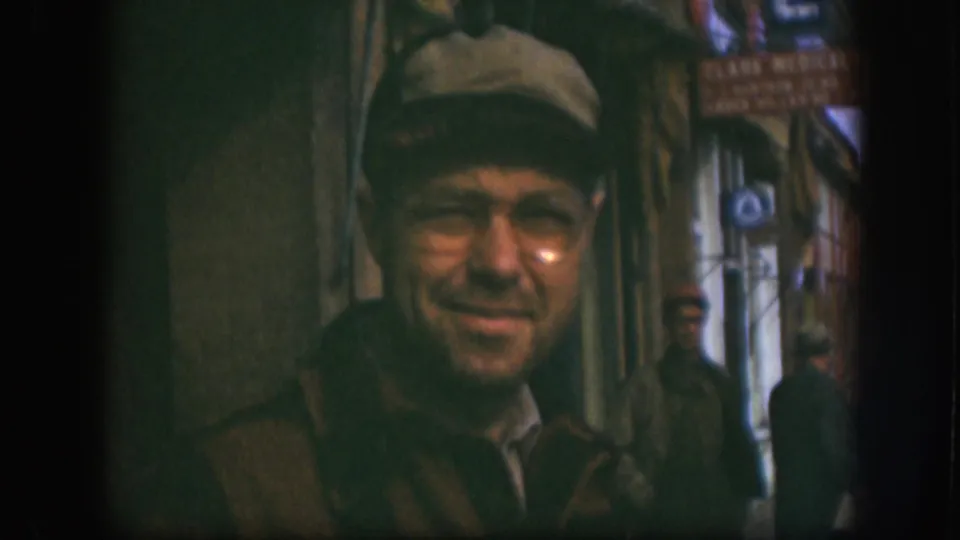 Close-up portrait of smiling man in newsboy cap and glasses at a Kansas small-town storefront, circa 1958; partial 'Clark Medical' sign at upper right of frame.