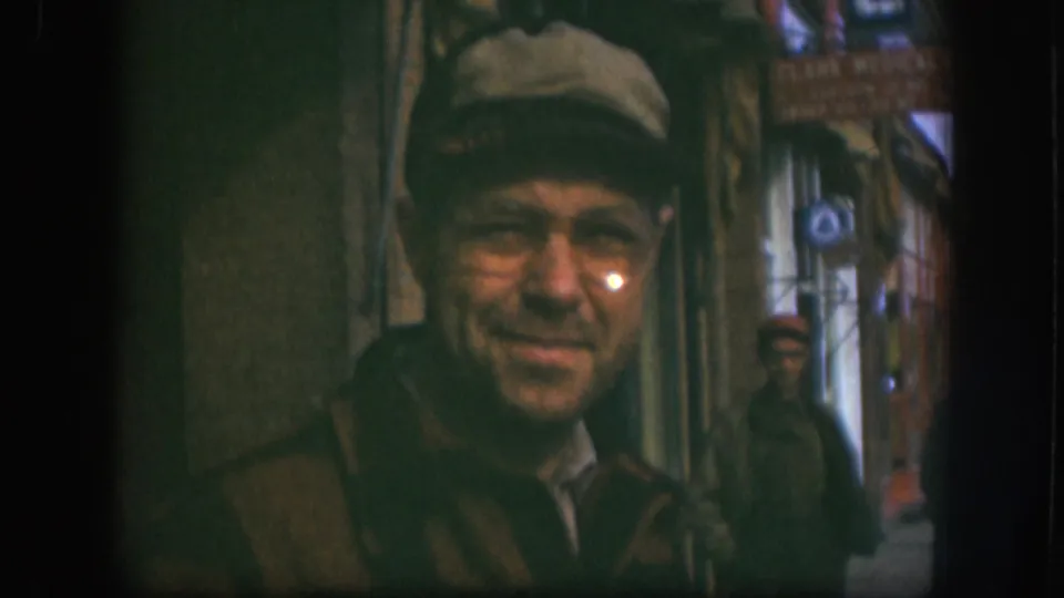 Man in newsboy cap and wire-rim glasses smiling toward the camera outside a Kansas small-town shop, circa 1958; 'Clark Medical Clinic' sign partially legible at upper right.