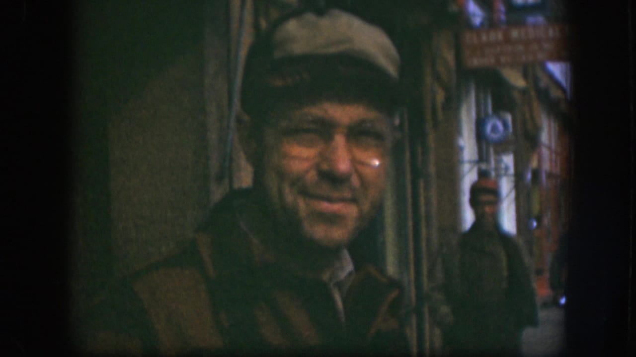 Candid 8mm close-up of an older man in flat cap and wire-rim glasses smirking at the camera on a Kansas street, circa 1958, with a storefront and CLARK MEDICAL sign behind.