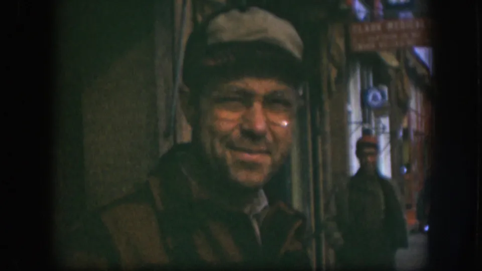 Candid 8mm close-up of an older man in flat cap and wire-rim glasses smirking at the camera on a Kansas street, circa 1958, with a storefront and CLARK MEDICAL sign behind.