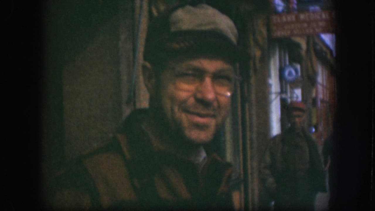 Older man in flat cap and glasses smiling, slightly shifted in frame; CLARK MEDICAL sign legible in upper-right background.