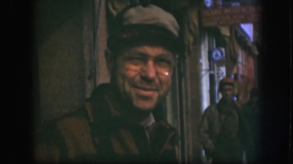 Close 8mm frame of man in flat cap and wire-rim glasses, face larger in shot, a second male figure visible against storefronts in the background, Kansas circa 1958.