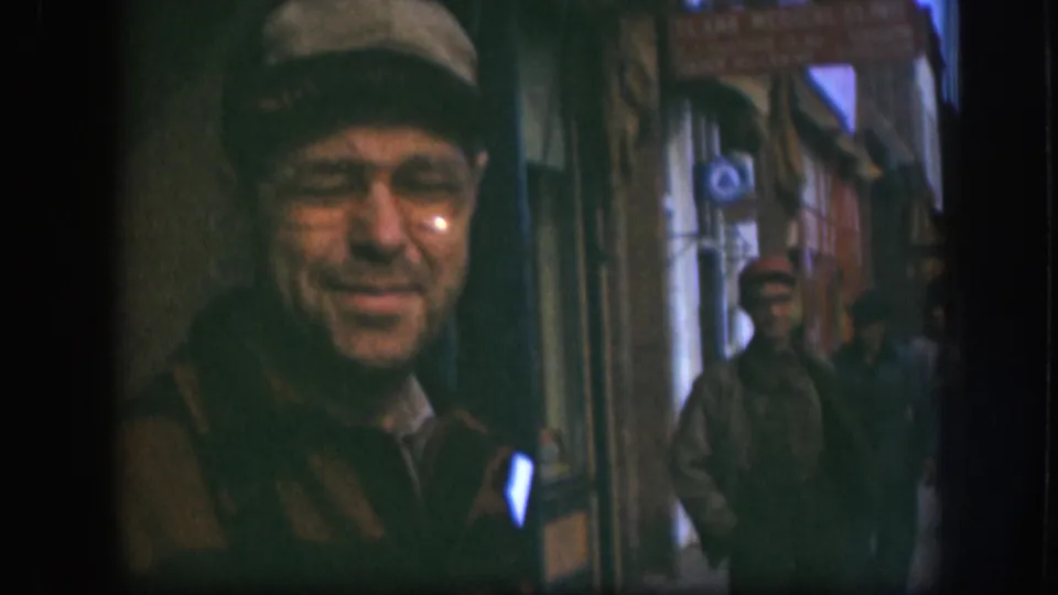 Final 8mm frame of Kansas street clip, circa 1958: man in flat cap and glasses with smirking expression, second male figure visible at the right edge of background storefronts.