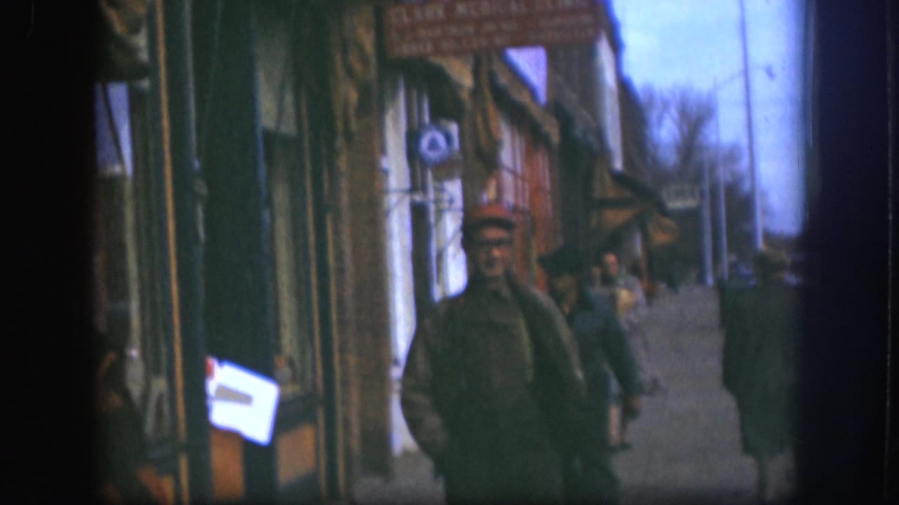 Man in red cap walking toward camera on a Kansas small-town commercial street, circa 1958; awning storefronts and other pedestrians visible behind him, 8mm color.