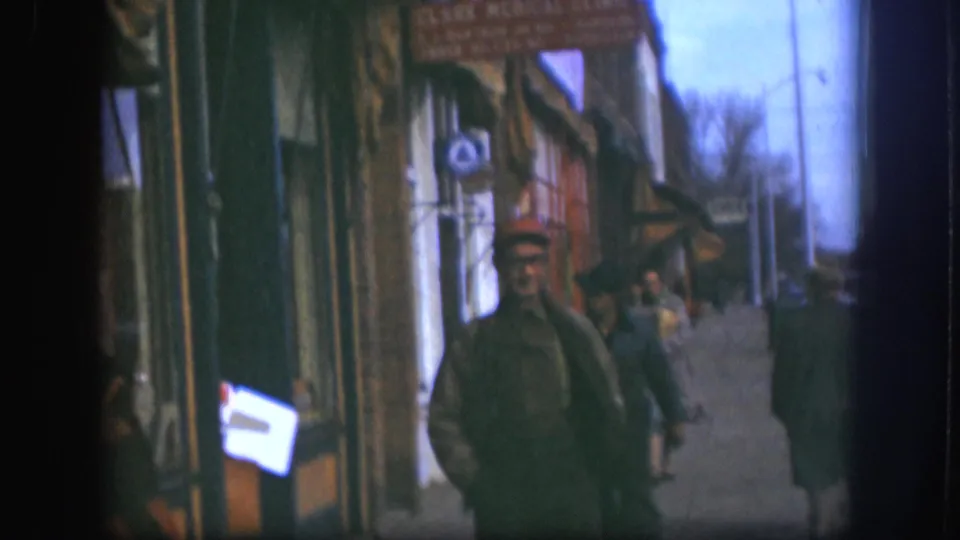Man in red cap walking toward camera on a Kansas small-town commercial street, circa 1958; awning storefronts and other pedestrians visible behind him, 8mm color.