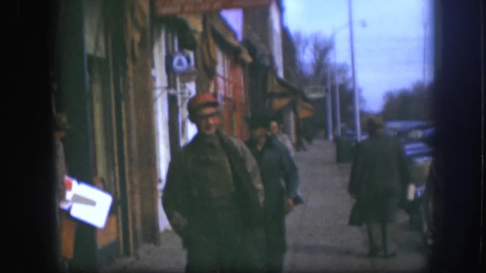 Man in red cap approaching camera on a Kansas commercial sidewalk circa 1958, with a woman pedestrian and period parked automobile visible behind him, 8mm color footage.