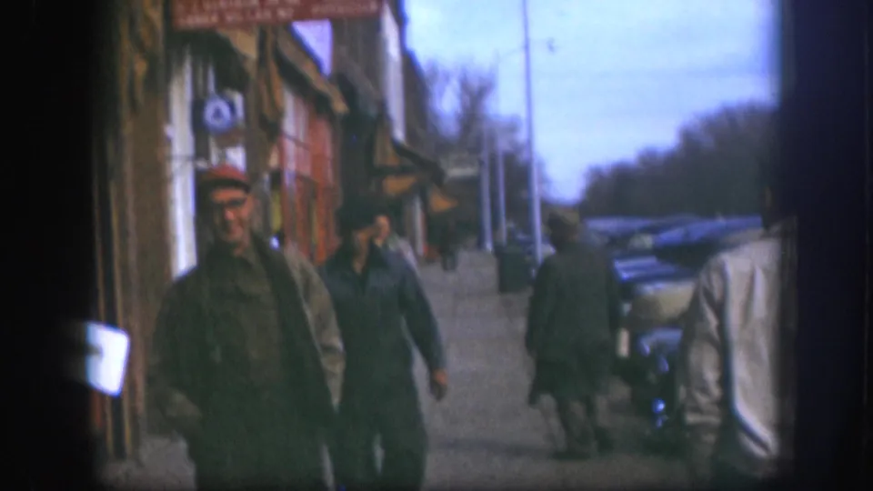 Man in red cap near camera on a Kansas small-town street circa 1958; late-1950s automobiles and awning-fronted shops visible along the block, 8mm color.