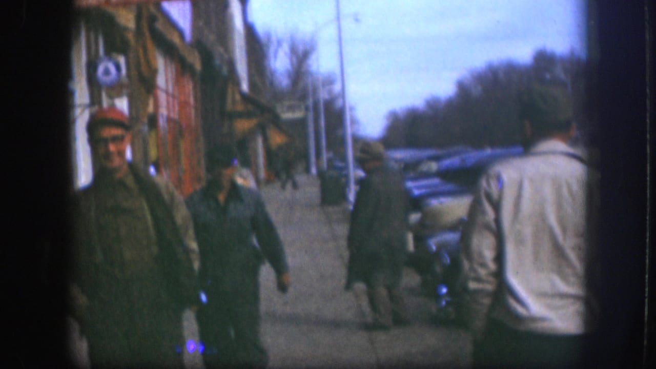 Two men on a sidewalk in what appears to be a small Kansas town, circa 1958; one faces the camera in a red cap, the other is seen from behind in a light jacket.