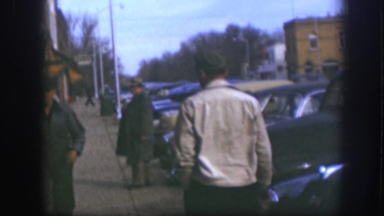 Two men walk along a sidewalk past parked classic sedans; one wears a light jacket, the other dark clothing.