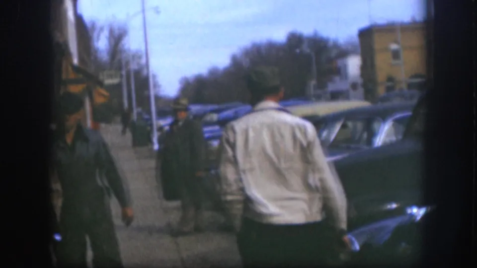 Pedestrians on a small-town main-street sidewalk, consistent with late-1950s Kansas; classic sedans parked along the curb and a brick building visible in the mid-background.