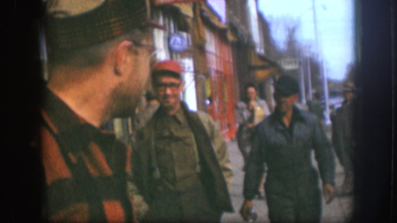 Man in plaid shirt and glasses in foreground; two men in work caps and jackets walking toward camera along a commercial sidewalk.