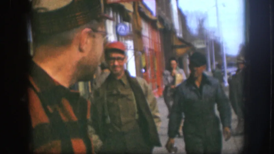 Close-up of a man in plaid flannel and glasses, with two men in caps and work jackets walking past storefronts on a Kansas street, circa 1958, 8mm.