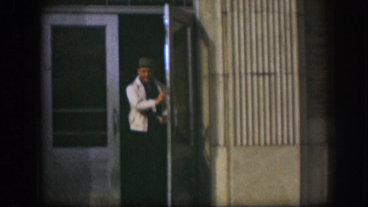 Man in white jacket and dark cap grasping the handle of a green glass-panel door on a corrugated metal building.