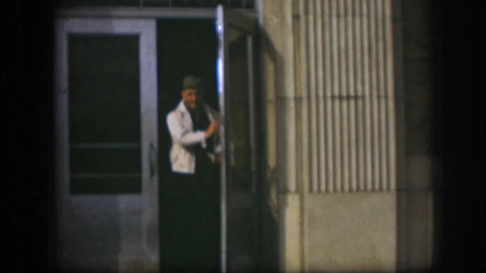 A man in a white jacket and dark cap reaches for a green glass-panel door on a corrugated metal storefront exterior, Kansas, circa 1958, 8mm.