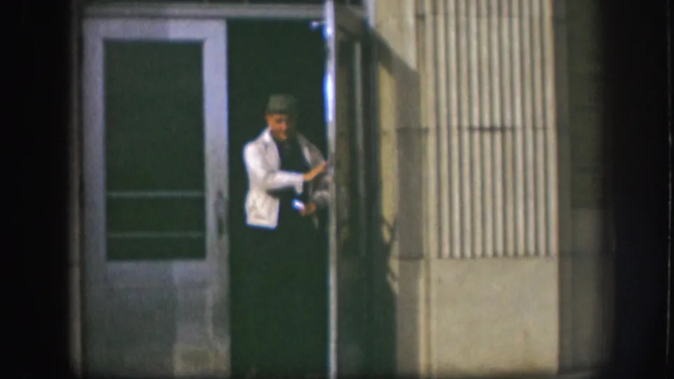A man in a white jacket and dark cap steps through a green door on a corrugated metal storefront, Kansas, circa 1958, 8mm footage.